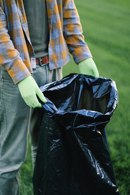 Close-up of a person wearing a green pair of gloves and a casual plaid shirt, standing outdoors on a grassy area, holding open a large black plastic rubbish bag with their hands. The bag appears to be
