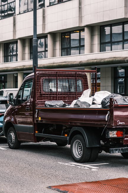 A small burgundy flatbed truck parked on an urban street in front of a modern, multi-storey office building with large glass windows and a light-colored stone facade. The truck's open bed is filled wi
